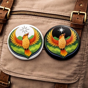 May include: Two round buttons with a bird design. One button has a white background, the other is black. The bird is orange and yellow with outstretched wings, a star above, and a green and blue landscape.