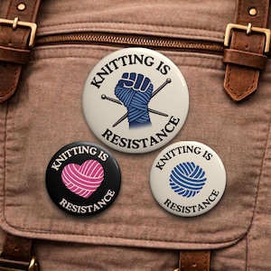 May include: Three round buttons with the text "Knitting is Resistance". One button features a blue fist wrapped in yarn and knitting needles. Another has a pink yarn heart. The third has a blue yarn ball.