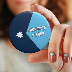 May include: A round button with a blue and light blue design, featuring the text "MINNESOTA STRONG" and a white star. The button is held in a person's hand.