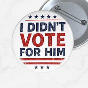 May include: A white button with the text "I DIDN'T VOTE FOR HIM" in red and blue. The button has a pin back and is surrounded by a silver metal rim. The design includes stars and stripes.