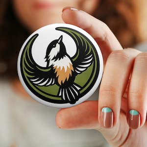 May include: A round, white button featuring a stylized bird with outstretched wings. The bird has black and white feathers, a tan chest, and is set against a green and black background. The button is held in a person's hand.