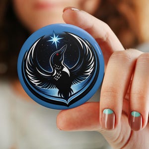 May include: A round, blue button featuring a stylized black bird with white wing details, a red eye, and a star. The bird is set against a starry night sky background. The button is held by a person.