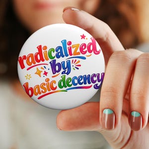 May include: A white button with the text "radicalized by basic decency" in a colorful, rainbow-like font. The button is held in a hand with painted nails. The button has a star and sparkle design.