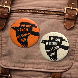 May include: Two round buttons with a raven graphic and the text "No one is illegal on stolen land." One button is orange, the other is off-white. The raven is black.