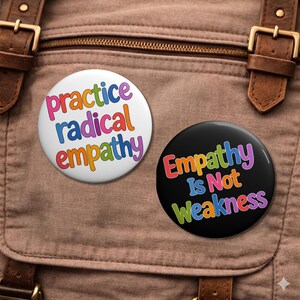 May include: Two round buttons with text on a brown backpack. One button is white with the words "practice radical empathy" in colorful letters. The other button is black with the words "Empathy Is Not Weakness" in colorful letters.