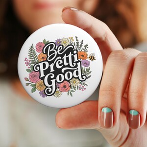 May include: A white round button with the words "Be Pretti Good" in black script, surrounded by colorful flowers and a small bee. The flowers include pink roses, orange poppies, and yellow daisies. A decorative item.