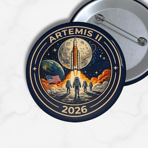 May include: A round, navy blue button with a space-themed design. The button features the text "ARTEMIS II" and "2026", along with an illustration of a rocket launching towards the moon, astronauts, the Earth, and the American flag.