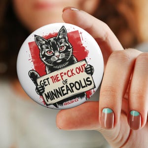 May include: A round, white button with a black cat illustration holding a sign that reads "GET THE F*CK OUT OF MINNEAPOLIS". The cat has red eyes and is set against a red background. The text "ABOLISH ICE" is at the bottom.