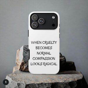 May include: White phone case with a black camera module and the text "WHEN CRUELTY BECOMES NORMAL COMPASSION LOOKS RADICAL" in black. The case is displayed on a rough, gray stone surface.
