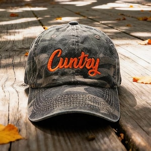 Könnte beinhalten: Eine Baseballkappe mit anthrazitfarbenem Tarnmuster, gebogenem Schirm und verstellbarem Riemen auf der Rückseite. Auf der Vorderseite ist das Wort "Cuntry" in einer leuchtend orangefarbenen Schreibschrift mit sichtbarer Stickstruktur aufgestickt.
