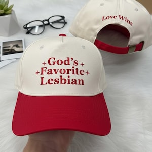 Könnte beinhalten: Cremefarbene Baseballkappe mit rotem Schirm. Auf der Vorderseite der Kappe steht in roter Schrift "God's Favorite Lesbian". Auf der Rückseite der Kappe steht in roter Schrift "Love Wins". Die Kappe hat einen roten, verstellbaren Riemen.