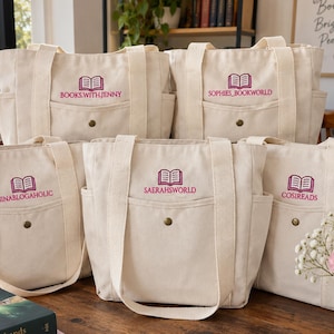 Könnte beinhalten: Fünf cremeweiße Canvas-Tragetaschen mit zwei stabilen Schulterriemen und einer Vordertasche mit Messingdruckknopf. Jede Tasche zeigt ein gesticktes, magentafarbenes Symbol eines offenen Buches über personalisiertem Text: BOOKS.WITH.JENNY, SOPHIES_BOOKWORLD, NINABLOGAHOLIC, SAERAHSWORLD und COSIREADS.