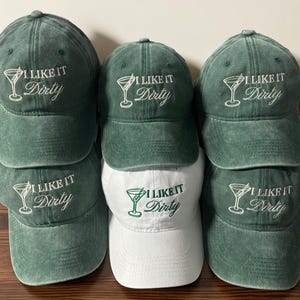 Puede incluir: Una pila de seis gorras de béisbol, cinco en verde apagado y una en blanco. Cada gorra presenta la frase bordada "I LIKE IT Dirty" con un diseño de copa de martini. Las gorras son de tela suave y texturizada.