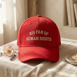 Könnte beinhalten: Rote Baseballkappe mit dem weißen Text "BIG FAN OF HUMAN RIGHTS". Die Kappe wird auf einer neutralfarbenen Oberfläche mit einer kleinen Kerze und Blumen in der Nähe präsentiert.