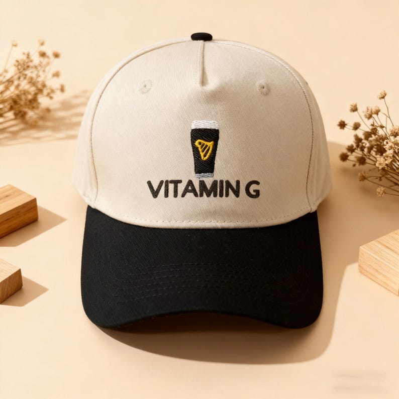 May include: A baseball cap with a cream-colored crown and a black brim. The cap features an embroidered design of a black pint glass with a yellow harp and the text "VITAMIN G" in black.