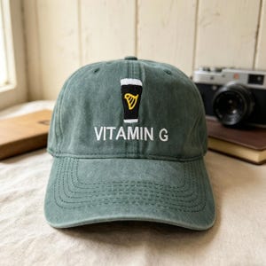 Könnte beinhalten: Eine grüne Baseballkappe mit dem gestickten Text "VITAMIN G" in Weiß. Oberhalb des Textes befindet sich eine schwarz-weiße Grafik eines Bierkrugs mit einem goldenen Harfensymbol. Die Kappe ist aus weichem, abgenutztem Material.