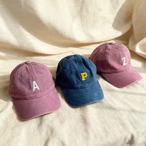 Könnte beinhalten: Drei Baseballkappen in Lila- und Blautönen werden gezeigt. Jede Kappe hat einen weißen oder gelben Buchstaben: "A", "P" und "Z". Die Kappen haben einen abgenutzten, Vintage-Look mit leicht verblichenem Aussehen. Die Kappen sind aus weichem Stoff.