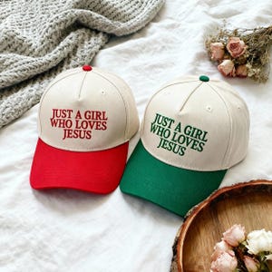 Könnte beinhalten: Zwei Baseballkappen mit dem Text "JUST A GIRL WHO LOVES JESUS". Eine Kappe hat einen roten Schirm und einen roten Knopf, die andere einen grünen Schirm und einen grünen Knopf. Die Kappen sind cremefarben.