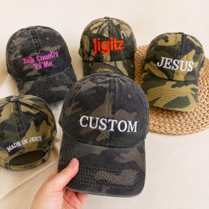 Könnte beinhalten: Eine Auswahl an Camouflage-Baseballkappen mit gestickten Texten in Kontrastfarben. Die Kappen zeigen Sätze wie "Talk Country To Me", "Jigitz", "JESUS" und "CUSTOM". Die Hüte haben ein verwaschenes, Vintage-Aussehen.