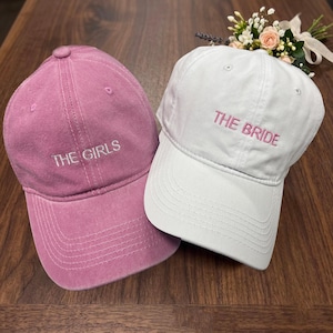 Könnte beinhalten: Zwei Baseballkappen, eine rosa und eine weiße, liegen auf einer Holzoberfläche. Auf der rosa Kappe steht "THE GIRLS" in Weiß gestickt, während auf der weißen Kappe "THE BRIDE" in Rosa steht. Hinter der weißen Kappe befindet sich ein kleines Blumengesteck.