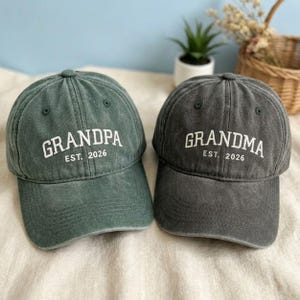 May include: Two baseball caps, one green and one grey, with white text. The green cap says "GRANDPA EST. 2026" and the grey cap says "GRANDMA EST. 2026". Both caps have a curved peak and an adjustable strap.