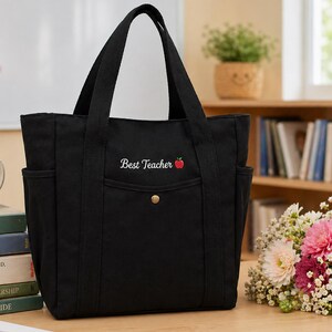 May include: Black canvas tote bag with two side pockets and sturdy handles. The front features the embroidered text "Best Teacher" with a red apple graphic. A gold-colored button secures the front pocket.