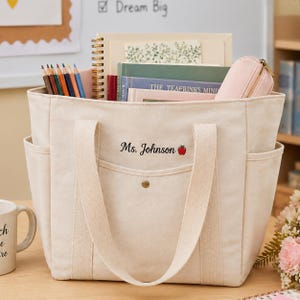 May include: A beige canvas tote bag with side pockets and long handles. The bag is filled with books, a pencil case, and a spiral notebook. The text "Ms. Johnson" and a red apple are embroidered on the front.