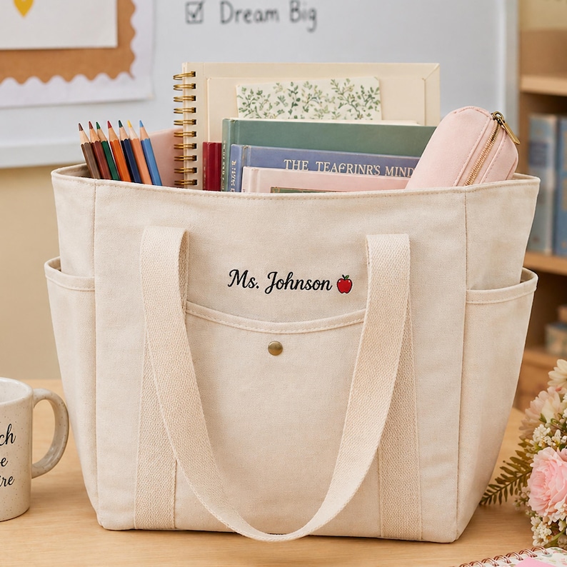 May include: A beige canvas tote bag with side pockets and long handles. The bag is filled with books, a pencil case, and a spiral notebook. The text "Ms. Johnson" and a red apple are embroidered on the front.