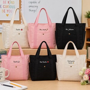 May include: Six canvas tote bags in cream, pink, and black, each with a front pocket and top handles. The bags are embroidered with teacher-themed text and graphics, including an apple and pencil. The bags are designed for teachers.