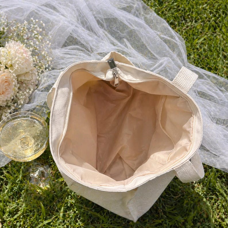 May include: Cream-colored tote bag with a zippered top, open to reveal a beige interior. The bag has sturdy handles and sits on green grass, with a glass of champagne and flowers nearby. A sheer white fabric is draped in the background.