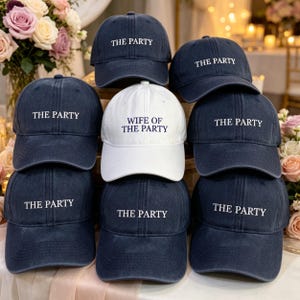 May include: A group of navy blue baseball caps, each embroidered with "THE PARTY" in white, and a single white cap with "WIFE OF THE PARTY". The caps are displayed together, with flowers visible in the background.