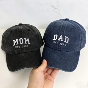 Pode incluir: Duas bonés de beisebol, uma preta e outra azul marinho. O boné preto tem a palavra "MOM" bordada em branco, com "EST 2026" abaixo. O boné azul marinho tem "DAD" bordado em branco, com "EST 2026" abaixo.