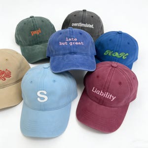 Può includere: Una collezione di cappellini da baseball in vari colori, tra cui verde, blu e bordeaux. Ogni cappellino presenta un ricamo con scritte come "papi", "late but great" e "Liability". I cappellini sono realizzati in un tessuto morbido e usurato.