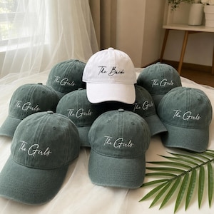 Könnte beinhalten: Eine Auswahl an Baseballkappen, darunter eine weiße Kappe mit "The Bride" und mehrere grüne Kappen mit "The Girls" in weißer Schrift. Die Kappen sind auf einer weichen, hellen Oberfläche angeordnet.
