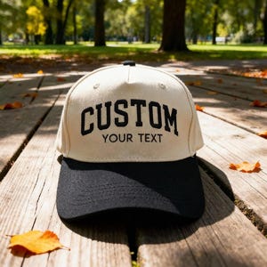 Könnte beinhalten: Beige und schwarze Baseballkappe mit dem Wort "CUSTOM" in fetten schwarzen Buchstaben und "YOUR TEXT" darunter. Die Kappe hat einen gebogenen Schirm und einen Knopf oben.
