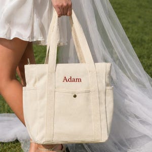 May include: Cream-colored canvas tote bag with the name "Adam" embroidered in red. The bag has two exterior side pockets, a front pocket, and sturdy handles. The bag is held by a person wearing a white dress.