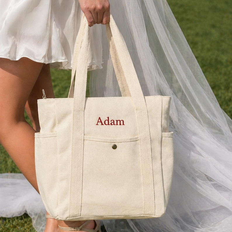 May include: Cream-colored canvas tote bag with the name "Adam" embroidered in red. The bag has two exterior side pockets, a front pocket, and sturdy handles. The bag is held by a person wearing a white dress.