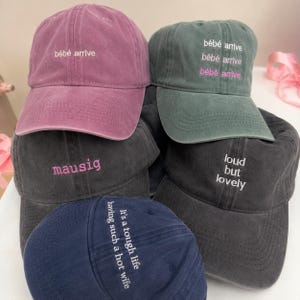 May include: A collection of five baseball caps in various colors including mauve, sage green, charcoal, and navy blue. Each cap features embroidered text on the front, including phrases like "bébé arrive", "mausig", "loud but lovely", and "It's a tough life having such a hot wife".
