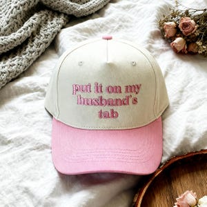 Puede incluir: Gorra de béisbol con corona color crema y visera rosa. La frase "put it on my husband's tab" está bordada en rosa en la parte delantera. La gorra está hecha de tela suave.