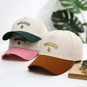 Könnte beinhalten: Drei Baseballkappen mit dem Wort "WINGLEADER" auf der Vorderseite gestickt. Die Kappen haben verschiedene Farbkombinationen: Beige mit grünem Schirm, Beige mit rosa Schirm und Beige mit braunem Schirm. Jede Kappe hat ein kleines Emblem unter dem Wort.