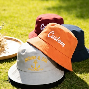 Könnte beinhalten: Eine Ansammlung von Fischerhüten in verschiedenen Farben, darunter Orange, Marineblau und Burgunderrot, liegt auf grünem Gras. Der orangefarbene Hut trägt den weißen Schriftzug "Custom". Der graue Hut hat "ANCHOR & SAIL" und einen gelben Anker.