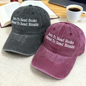 Könnte beinhalten: Zwei Baseballkappen, eine anthrazitgraue und eine weinrote, werden gezeigt. Beide Kappen haben den weißen Schriftzug "Born To Read Books, Forced To Read Emails". Die Kappen sind aus einem weichen, abgenutzten Material.