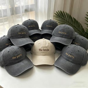May include: A collection of baseball caps. Most are dark gray with "the party" embroidered in gold thread. One cap is beige with "the bride" embroidered in gold. The caps have a vintage, worn look.