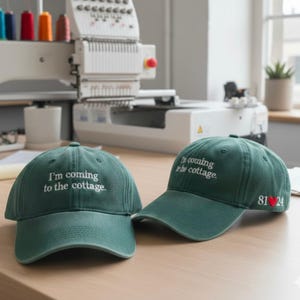 Könnte beinhalten: Zwei grüne Baseballkappen mit weiß gesticktem Text "I'm coming to the cottage." Eine Kappe hat die Stickerei "81 ❤️ 24" an der Seite. Die Kappen liegen auf einer Holzoberfläche.