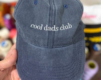 May include: A navy blue baseball cap with a distressed, washed-out finish. The front of the cap features the embroidered white text "cool dads club" in a lowercase serif font. The hat includes a curved brim and matching navy blue stitching.