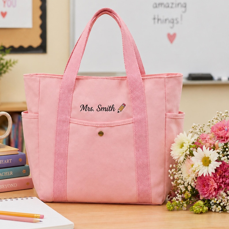 May include: A pink canvas tote bag with two handles and side pockets. The bag has the text "Mrs. Smith" and a pencil graphic embroidered on the front. A bouquet of flowers sits to the right of the bag.