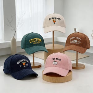 Peut inclure: Cinq casquettes de baseball en velours côtelé de différentes couleurs : bleu marine, vert, beige, marron et rose. Chaque casquette présente des textes et des motifs brodés, dont "Custom Design", "Be Yourself", "Camp Emily" et "I'm fine."