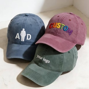 May include: Three baseball caps in blue, burgundy, and green. The blue cap features "A D" with a figure. The burgundy cap has "CUSTOM" embroidered in rainbow colours. The green cap displays "your logo". All caps have a curved peak.