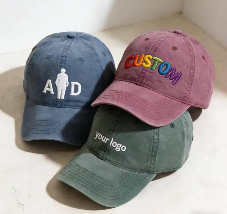 May include: Three baseball caps in blue, burgundy, and green. The blue cap features "A D" with a figure. The burgundy cap has "CUSTOM" embroidered in rainbow colours. The green cap displays "your logo". All caps have a curved peak.