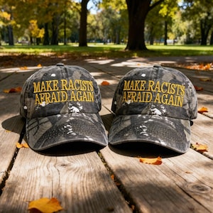 Könnte beinhalten: Zwei Camouflage-Baseballkappen mit dem gestickten Text "MAKE RACISTS AFRAID AGAIN" in Gelb. Die Hüte werden auf einer Holzoberfläche mit Herbstlaub vor einem verschwommenen Hintergrund im Freien gezeigt.
